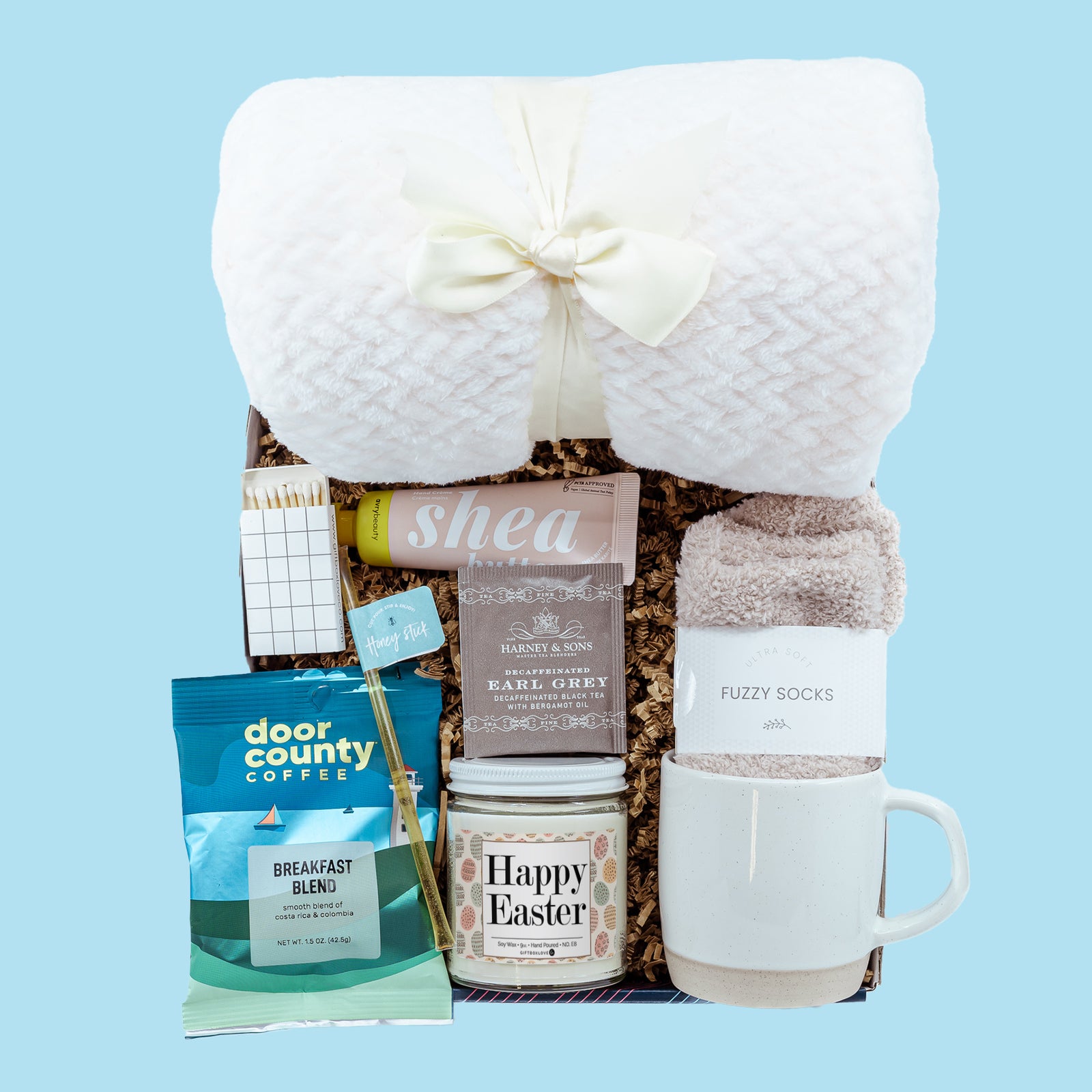 Easter Snuggle & Sip Gift Box featuring a soft blanket, coffee mug, 9 oz candle, socks, coffee, tea, honey, hand cream, matches, and a handwritten card.