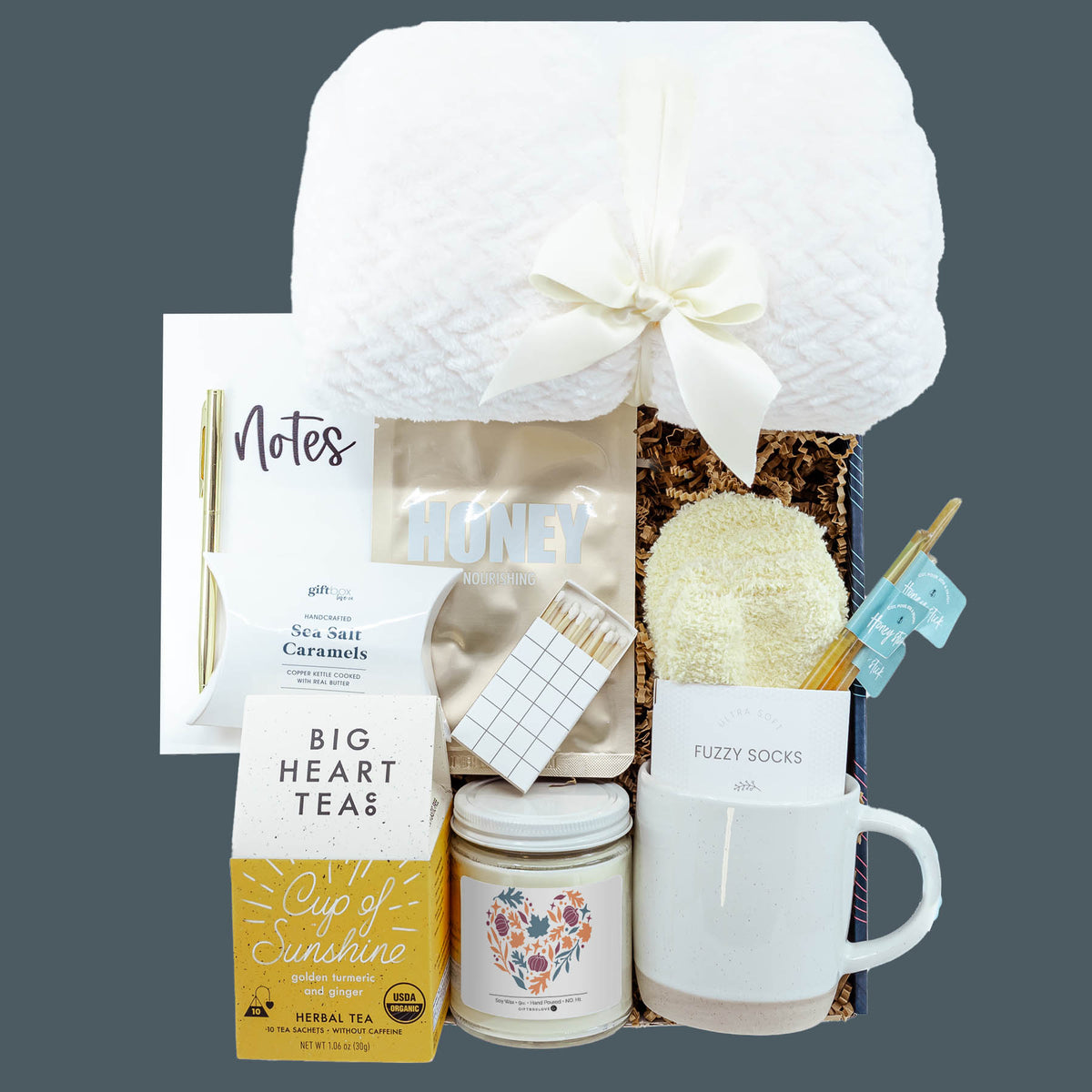 Harvest Hugs Gift Box with plush blanket, stoneware mug, herbal tea, autumn soy candle, fuzzy socks, notebook, honey face mask, sea salt caramels, honey sticks, and decorative matches.