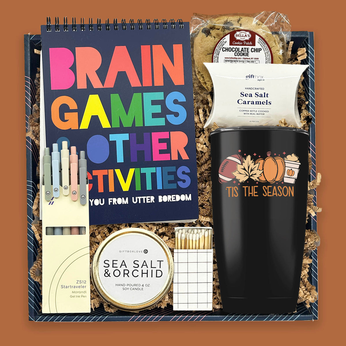 The Autumn Escape Gift Box with brain games book, gel pens, fall tumbler, chocolate chip cookie, sea salt caramels, soy candle, and decorative matches.
