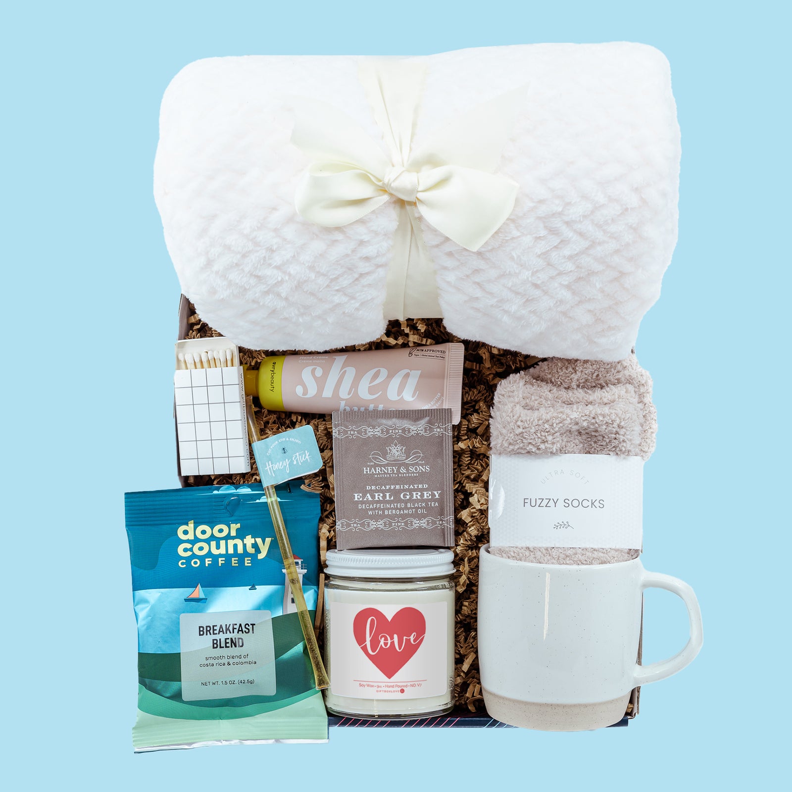 Valentine’s Snuggle & Sip Gift Box featuring a soft blanket, coffee mug, 9 oz candle, cozy socks, coffee, tea, honey stick, hand cream, matches, and a handwritten card.